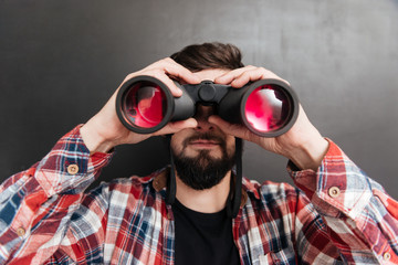 Serious bearded young man standing and looking through binoculars