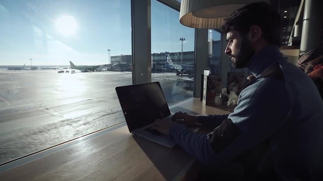 
  A guy   writes a text in the  laptop near the window in a cafe at the airport.