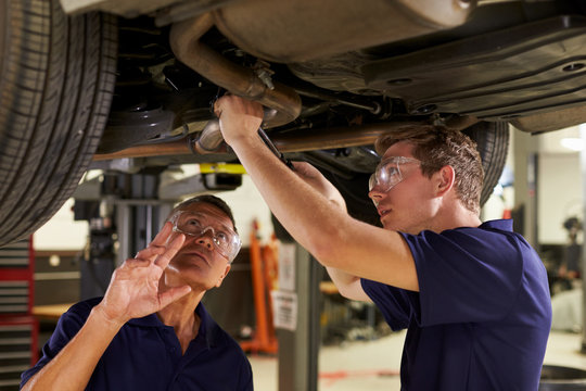 Mechanic And Male Trainee Working Underneath Car Together