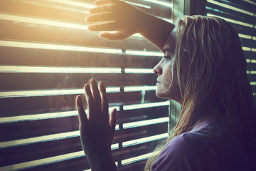 Sad and lonely blonde woman with wet hair looking through window blinds into the sunlight