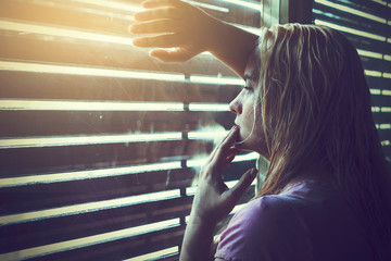 Sad and lonely blonde woman with wet hair looking through window blinds into the sunlight