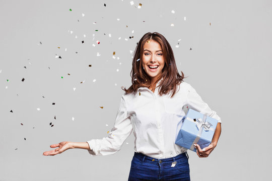 Beautiful Happy Woman With Gift Box At Celebration Party With Confetti Falling Everywhere On Her. Birthday Or New Year Eve Celebrating Concept