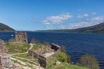 Naklejka premium Loch Ness, Szkocja - 2 czerwca 2012: Ujęcie szerszego ciemnoniebieskiego jeziora Loch Ness z częścią ruin zamku Urquhart na pierwszym planie. Zielone okoliczne wzgórza. Jasnoniebieskie niebo.