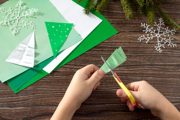 The child makes paper fir tree. Scissors, paper on a wooden tabl