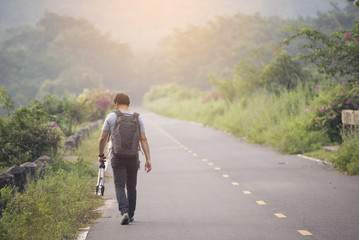 Obraz premium Lonely man holding a camera and a tripod,walking on the road ,sunsets and mountains as a backdrop.