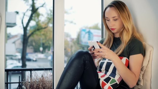 Absorbed girl sitting by the window and typing message on smartphone
