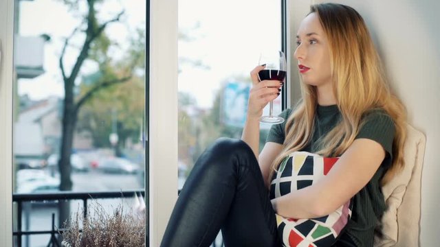 Sad girl drinking wine by the window and thinking about her problems
