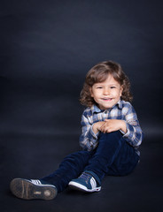 Cute curly baby with a charming smile and white teeth in fashionable plaid shirt sitting on a dark background.