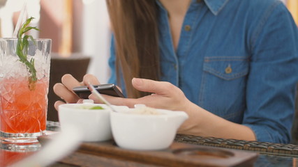 Brunette girl uses smart phone while sitting outside in a cafe, drinking a cool drink from a straw tube. 4k,