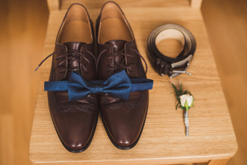 Men's fashion. Top view of groom's leather shoes, bow tie, belt and boutonniere on wooden chair.