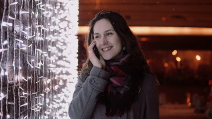 young attractive woman talking on the phone in the falling snow at Christmas night standing near lights wall,