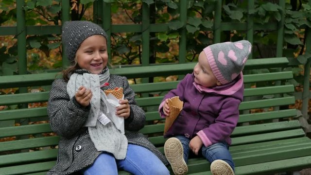 Two sisters sitting on the bench and eating ice-cream. Little girls sit in the park and eating ice cream. Children funning