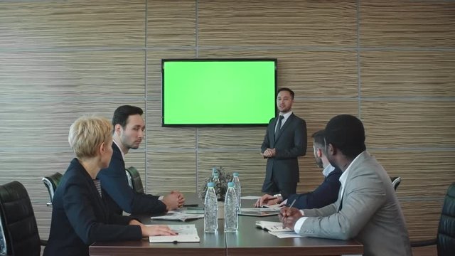 Lockdown of Asian businessman standing before LED TV screen with green chroma key background and showing presentation to colleagues sitting at table in conference room
