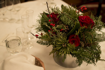 Elegant Christmas Centerpiece with Holly Mistletoe and red Roses on Table with white tablecloth, formal place setting with water, champagne flutes and bread basket