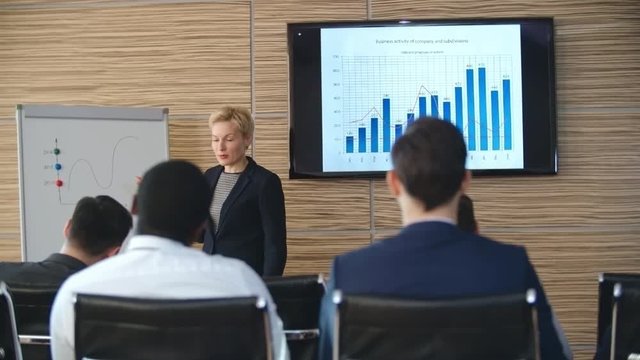 PAN of blond businesswoman standing before whiteboard and drawing something on finance chart