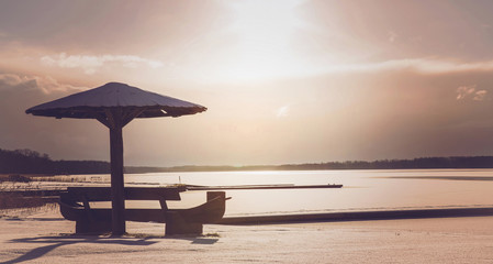 Wooden bench with umbrella on lake cowered with snow at beautiful sunset in winter evening. Focus point on the bench and umbrella.