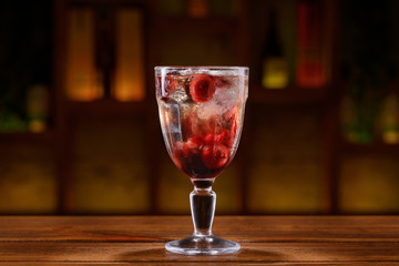 Cocktail with alcohol, cherries and ice cubes on a wooden table with blurred background of bottles and light. 