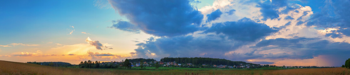 Sunset in the countryside. Dramatic sky with clouds, field and rural houses on a background of trees. Panoramic shot.