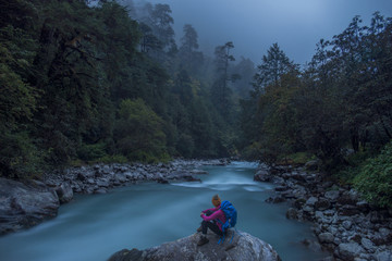 A woman takes a break from the trail and sits beside the Langtang Khola near the little village of Riverside on a misty evening, Nepal, Himalayas