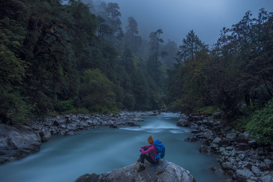 A woman takes a break from the trail and sits beside the Langtang Khola near the little village of Riverside on a misty evening, Nepal, Himalayas