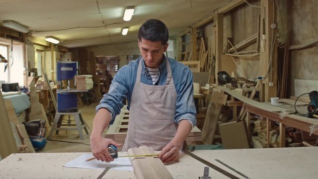PAN of woodworker standing in workshop and measuring width and height of board lying on workbench 