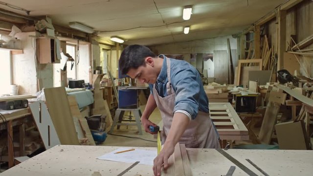 PAN of male carpenter checking width and height of wood board with tape measurer