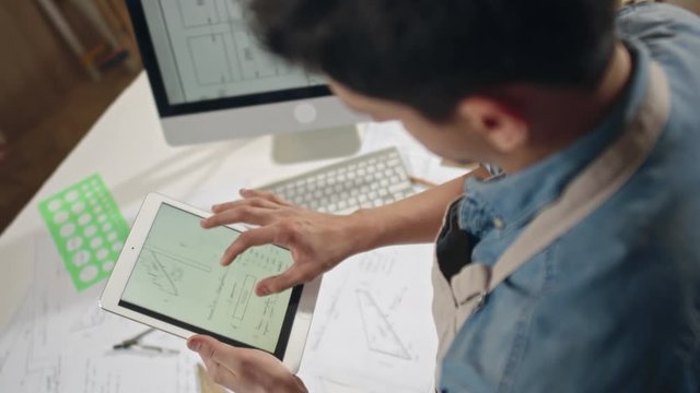High angle over the shoulder shot of male artisan looking at blueprints on tablet and computer screen and checking measurements 