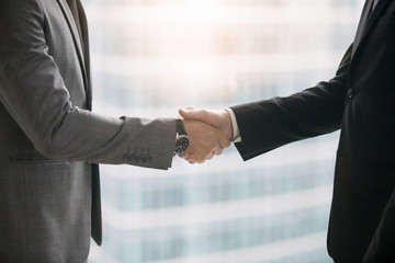 Close up of hands of two business partners making agreement, taking a big deal and shaking hands in meeting room at office. Urban style glass office building in big city on the background. Side view