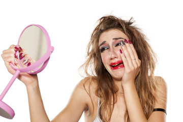 crying young woman with bad makeup and messy hair looking at herself in the mirror