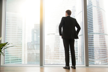 Fototapeta premium Full length back view of successful businessman in suit standing in office with hands on his waist, CEO looking through window at big city buildings, planning new project, waiting for meeting to start