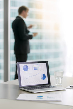 Company Leader In Classical Formal Suit Standing, Looking At Window And Drinking Coffee. Laptop With Rising Graphs On Office Table On Foreground, Focus On The Screen. Business Success Concept