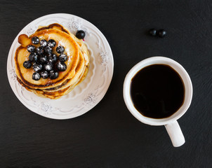 Homemade pancakes and black currant, vintage white crockery, cup of coffee, black stone background, top view.