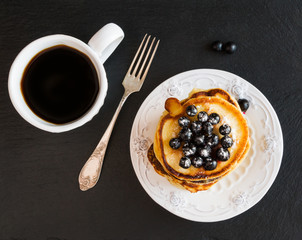 Homemade pancakes and black currant, vintage white crockery, cup of coffee, black stone background, top view.