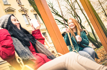 Joyful girlfriends having fun on swing game at park in autumn season - Friendship concept with happy girls sharing winter time together - Warm filter and sun flare halo with focus on blond young woman