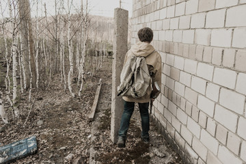 young boy stands near the abandoned building. wandering boy. man in a protective cloak with a hood. Post apocalypse. traveling on foot in a post-apocalyptic world.