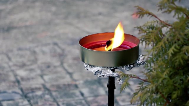 Flammenschale mit heller gelb-orangener Flamme und lichtreflektierendem Wachs / Nahaufnahme einer Flammschale mit rotem Wachs und Lichtreflexen auf der Wachsoberfl&auml;che und dem Metall der Schale.