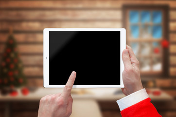 Tablet in Santa Claus hand with blank screen for mockup. Santa Claus home with Christmas tree in...
