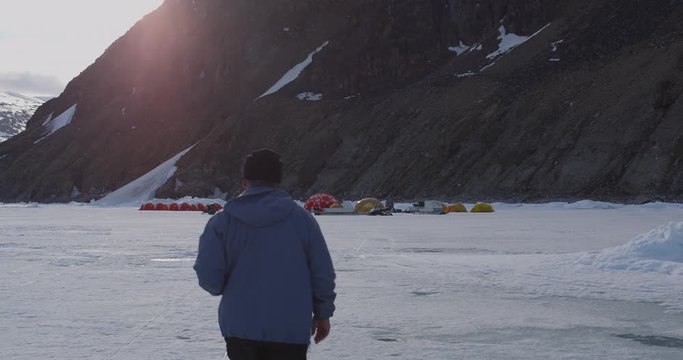 Tourist walks towards tents on sea ice below mountain at sunset