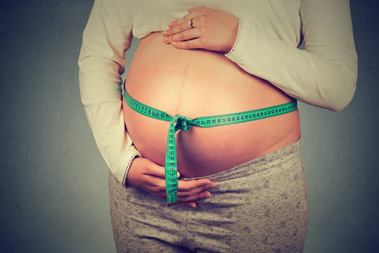Pregnant woman measuring belly standing against gray background