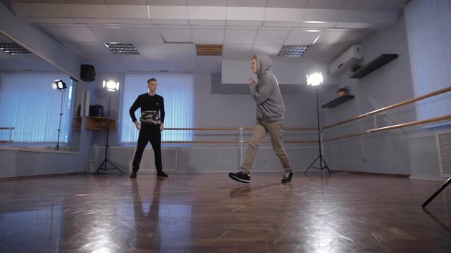 Two young hip-hop dancers participating in dance battle. Teenager in black trousers and pullover watching his friend performing urban dance in the classroom.
