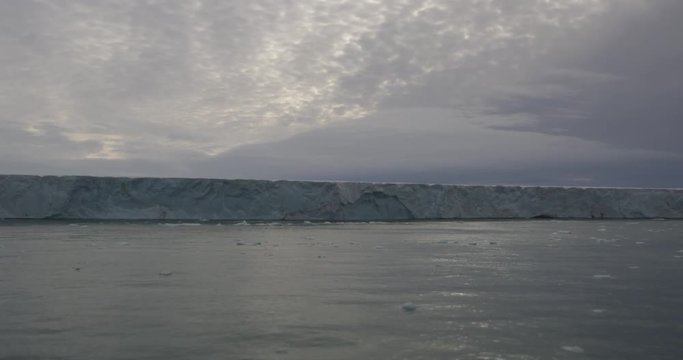 Passing large glacier cliff on calm arctic ocean at dusk
