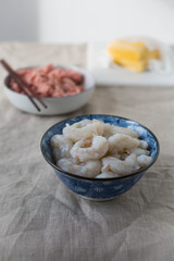 A blue bowl with raw shrimp. Ground pork and wonton wrappers in the background. 