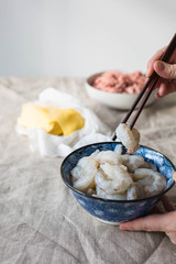 Raw shrimp in a blue bowl. Hands holding a pair of chopsticks. Wonton wrappers and ground pork in the background. 