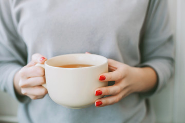 Young woman with bright red manicure and grey sweater holding a big white mug of tea with lemon