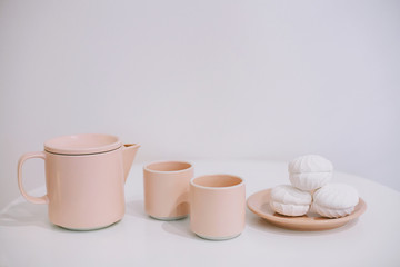 White table with a pink teapot and mugs and some tasty marshmallows on a plate in a white room
