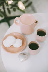 White table with a pink teapot and mugs and some tasty marshmallows on a plate in a white room near the christmas tree