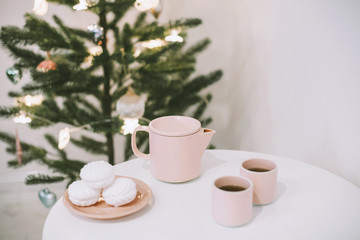 White table with a pink teapot and mugs and some tasty marshmallows on a plate in a white room near the christmas tree