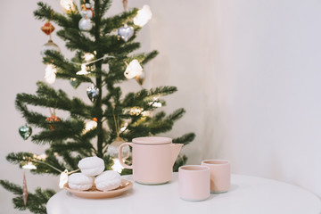 White table with a pink teapot and mugs and some tasty marshmallows on a plate in a white room near the christmas tree