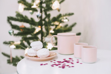 Obraz na płótnie Canvas White table with a pink teapot and mugs and some tasty marshmallows on a plate in a white room near the christmas tree