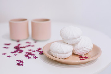 White table with a pink teapot and mugs and some tasty marshmallows on a plate in a white room